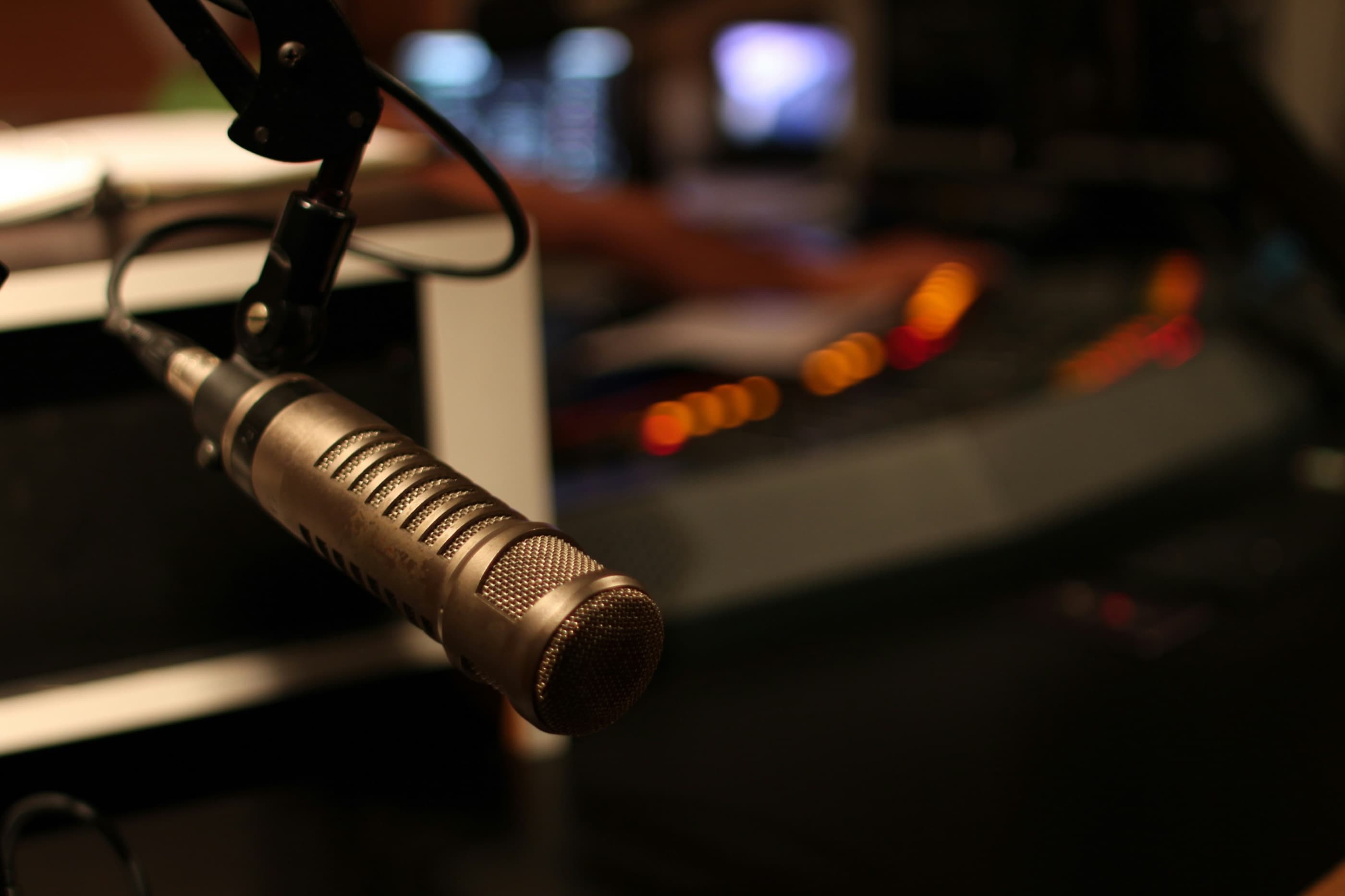 A worn broadcast microphone on an arm in a studio, with warm desk lights blurred behind it